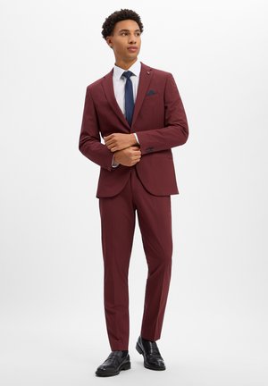 Jonge man draagt een bordeaux pak, wit overhemd, donkerblauwe stropdas, zwarte schoenen en stelt zijn colbertje bij tegen een effen witte achtergrond.