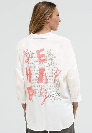 Woman with light brown hair wearing a white long-sleeve shirt with colorful text and drink illustrations on the back.