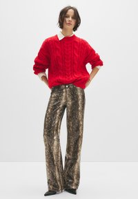 Maglione rosso a trecce con scollatura a girocollo, abbinato a pantaloni larghi con stampa serpente metallica oro e nera. Scarpe nere a punta.