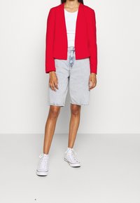 Punainen cropped-bleiseri, valkoinen tiukka t-paita, vaaleat farkkushortsit ja valkoiset korkeat tennarit. Silkkinen kangas, muotoiltu design, neutraalit sävyt.