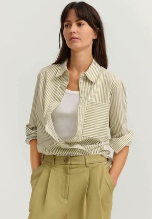 Frau mit langen dunklen Haaren, trägt ein beigefarbenes und weiß gestreiftes Hemd über einem weißen Trägertop, kombiniert mit hoch taillierten beigen Hosen, Hände in den Taschen.