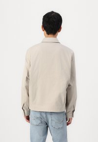 Beige jacket with a pointed collar, long sleeves, and cuff detailing, worn over light blue jeans. Smooth texture, casual fit.