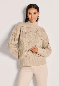 Frau in beigem Kabelstrickpullover mit Blättermustern und hellen Hosen, die vor einem schlichten hellen Hintergrund steht.
