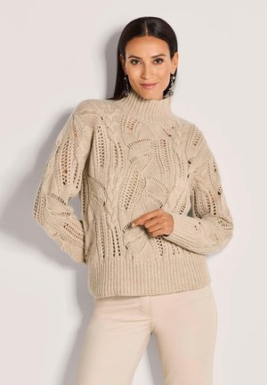 Vrouw in een beige kabelgebreid sweater met loofpatronen en lichte broek, staand tegen een effen lichte achtergrond.