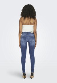 Femme aux cheveux bouclés portant un top caraco blanc, un jean skinny bleu avec des poches arrière et des chaussures à talons noires, debout de dos.