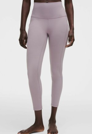 Zbliżenie osoby noszącej dopasowane do kostek legginsy w kolorze mauve, stojącej boso na jednolitym białym tle.