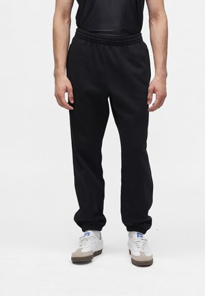 Mann trägt schwarze lässige Joggerhose, ein schwarzes ärmelloses Top und weiße Sneaker mit blauem Logo, steht vor weißem Hintergrund.
