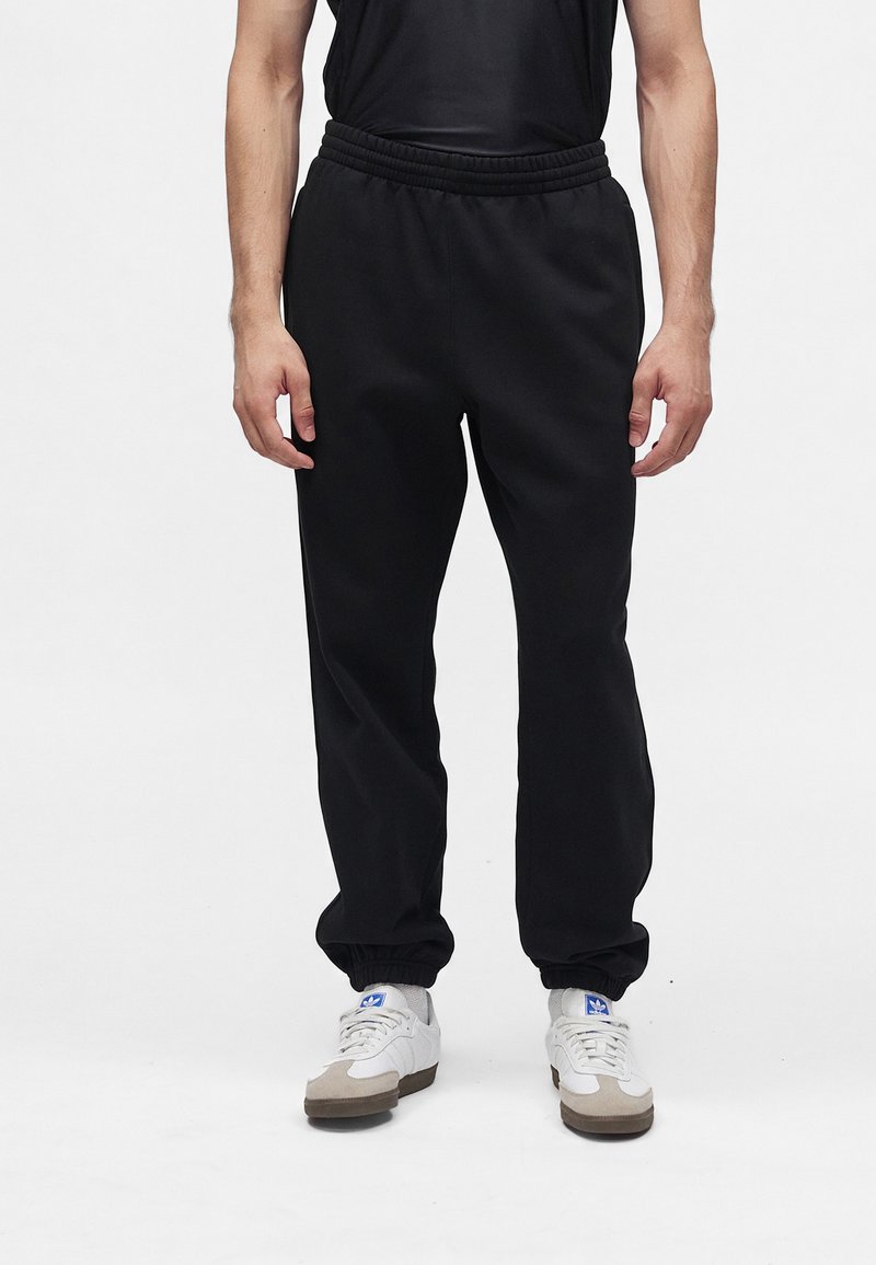 Άνδρας που φοράει μαύρο casual παντελόνι jogger, μαύρο αμάνικο τοπ και λευκά αθλητικά παπούτσια με μπλε λογότυπο, στέκεται σε λευκό φόντο.
