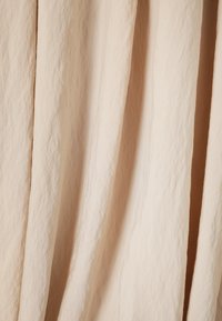 Tessuto beige chiaro con una superficie morbida e texture, caratterizzato da pieghe sottili e drappeggi, che creano linee fluide.