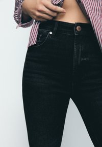 Črne visokorasle kavbojke s ožjim krojem. Lastnosti vključujejo sprednje zapiranje na gumb, zanke za pas ter rahlo teksturirano površino.