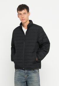 Jachetă neagră izolată cu cusături verticale, guler înalt și închidere cu fermoar; are buzunare laterale; asociată cu blugi din denim deschis.