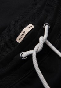 Zwarte trainingsbroek met een elastische tailleband, witte trekkoorden en een klein merklabel met "ombre" en maat M. Zachte textuur zichtbaar.