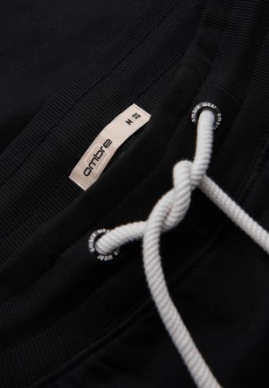 Pantalons de survêtement noirs avec une taille élastique, un cordon blanc et une petite étiquette de marque affichant "ombre" et taille M. Texture douce visible.