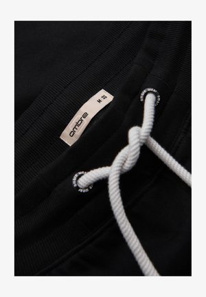 Pantalones de chándal negros con cinturilla elástica, cordón blanco y pequeña etiqueta de marca que muestra "ombre" y talla M. Textura suave visible.