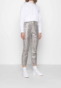 Metalliska silverfärgade capribyxor med ett texturerat reptilmönster, i kombination med en vit huvtröja och vita sneakers, mot en enfärgad bakgrund.