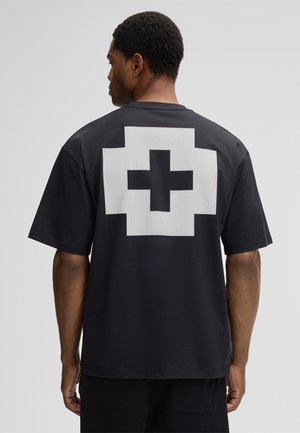 Man facing backward wearing a black t-shirt with a large white pixelated cross design on the back, against a plain gray background.