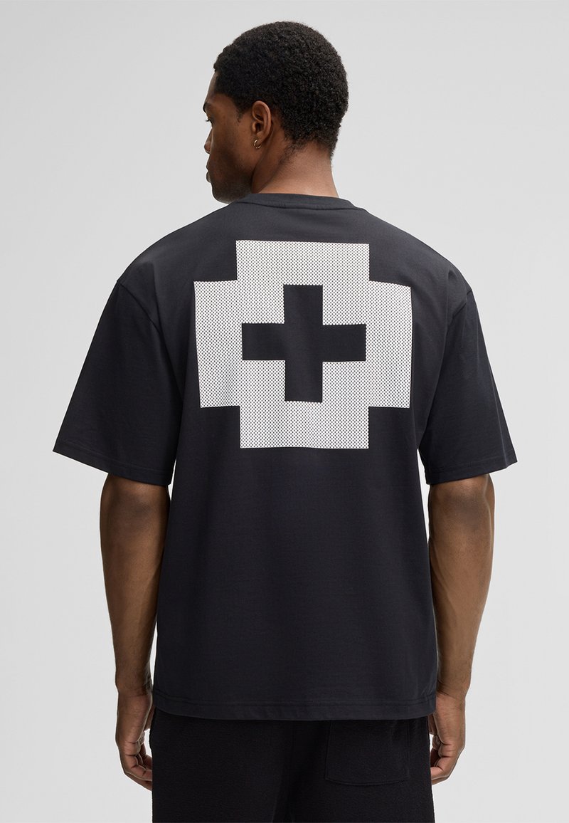 Mann mit dem Rücken zur Kamera, der ein schwarzes T-Shirt mit einem großen, weißen, pixelierten Kreuzdesign auf dem Rücken trägt, vor einem schlichten grauen Hintergrund.