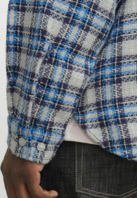 Woven plaid shirt in gray, blue, and black with a textured finish, featuring buttoned cuffs and a side vent detail, layered over dark jeans.