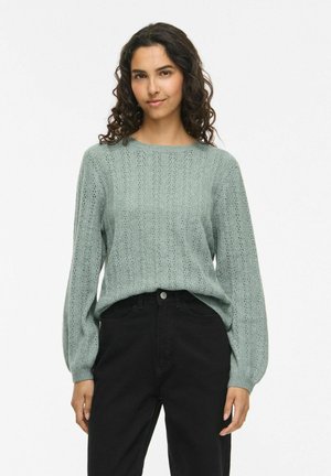 Femme aux cheveux bouclés foncés porte un pull en maille vert clair avec des manches ballon et un pantalon noir taille haute sur un fond uni.
