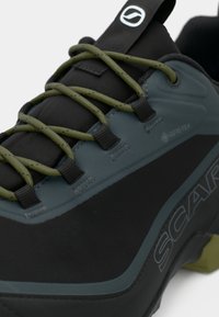 Zapato de senderismo negro y gris con parte superior de Gore-Tex, malla texturizada, cordones verdes y un logotipo prominente en la lengüeta y el costado.
