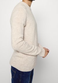 Pull beige en tricot doux, col rond, manches longues, avec des poignets et un ourlet côtelés, associé à un jean bleu.