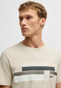 Camiseta de algodón beige con diseño gráfico abstracto que presenta rectángulos en negro, blanco y gris, patrones texturizados y escote redondo.