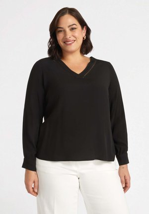 Blusa nera a maniche lunghe con scollo a V, caratterizzata da un motivo testurizzato vicino al collo e una vestibilità ampia, abbinata a pantaloni bianchi.