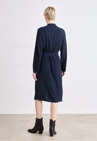 Navy blue dress with long sleeves, a belted waist, and a rounded hem, paired with black heeled ankle boots. Smooth fabric texture.