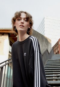 Felpa nera con design a tre strisce bianche sulle maniche e piccolo logo Adidas bianco sul petto. Vestibilità casual e rilassata. Il modello posa sulle scale.