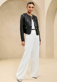 Melna ādas īsā jakā ar apaļu kakla izgriezumu un zelta pogu aizdari, kopā ar baltām platām biksēm un sudraba aksesuāru apaviem.