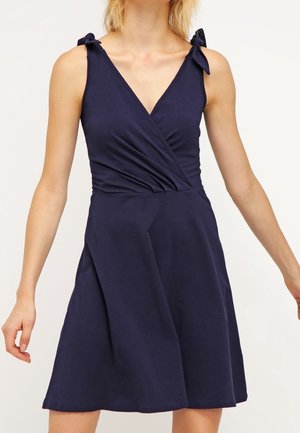 Femme portant une robe portefeuille sans manches bleu marine avec des bretelles nouées aux épaules et une jupe évasée sur fond uni.