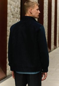 Chaqueta de ante azul marino con un collar alto, que presenta una textura suave y un diseño minimalista. El modelo se encuentra de pie frente a una pared de ladrillos texturizada.