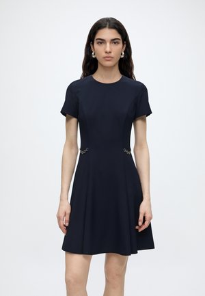 Frau mit langen dunklen Haaren, die ein marineblaues kurzärmeliges Kleid mit silbernen Kettendetails an der Taille trägt, steht vor einem schlichten weißen Hintergrund.