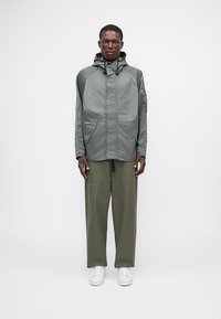 Ελαφρύ, ανθεκτικό στο νερό γκρι μπουφάν με κουκούλα, δύο μπροστινές τσέπες και ρυθμιζόμενα κορδόνια, συνδυασμένο με ελαιοπράσινα παντελόνια και λευκά αθλητικά παπούτσια.