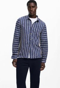 Prúžkovaná košeľa na gombíky v námorníckej modrej, bielej a zelenej farbe. Má golier a dlhé rukávy, vyrobená z ľahkej látky s voľným strihom.