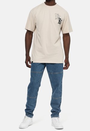 Camiseta de algodón beige con un gráfico negro en el pecho izquierdo, combinada con pantalones cargo de mezclilla azul con múltiples bolsillos y un corte holgado.