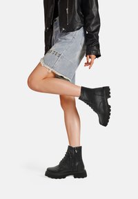 Stivaletti neri in pelle con lacci e suola spessa, caratterizzati da cerniere laterali e dettagli texture, abbinati a una gonna di jeans.