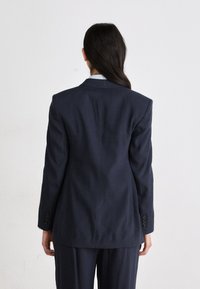 Blazer blu navy con forma strutturata, chiusura a un petto e quattro bottoni decorativi sulle maniche. Tessuto testurizzato e rever definiti.