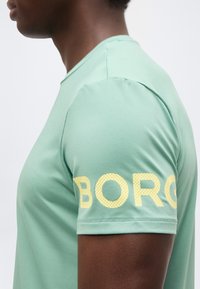 Πλευρική όψη ατόμου που φοράει μπλουζάκι σε μέντα πράσινο χρώμα με μεγάλα κίτρινα γράμματα «BORG» στο μανίκι, μπροστά σε μονόχρωμο φόντο.