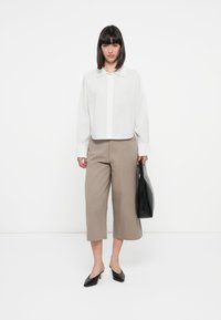 Weißes Kragenhemd, beige verkürzte weite Hosen, schwarze Absatzmules, hält eine schwarze Tragetasche, vor einem schlichten weißen Hintergrund.
