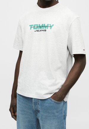 Mężczyzna noszący jasnoszarą koszulkę Tommy Jeans z turkusowym i czarnym napisem, w zestawieniu z niebieskimi dżinsami, ręka w kieszeni na białym tle.