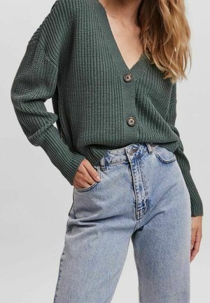 Kobieta ubrana w zielony dziergany kardigan z dwoma guzikami oraz jasnoniebieskie wysokowalne dżinsy, z ręką w kieszeni.