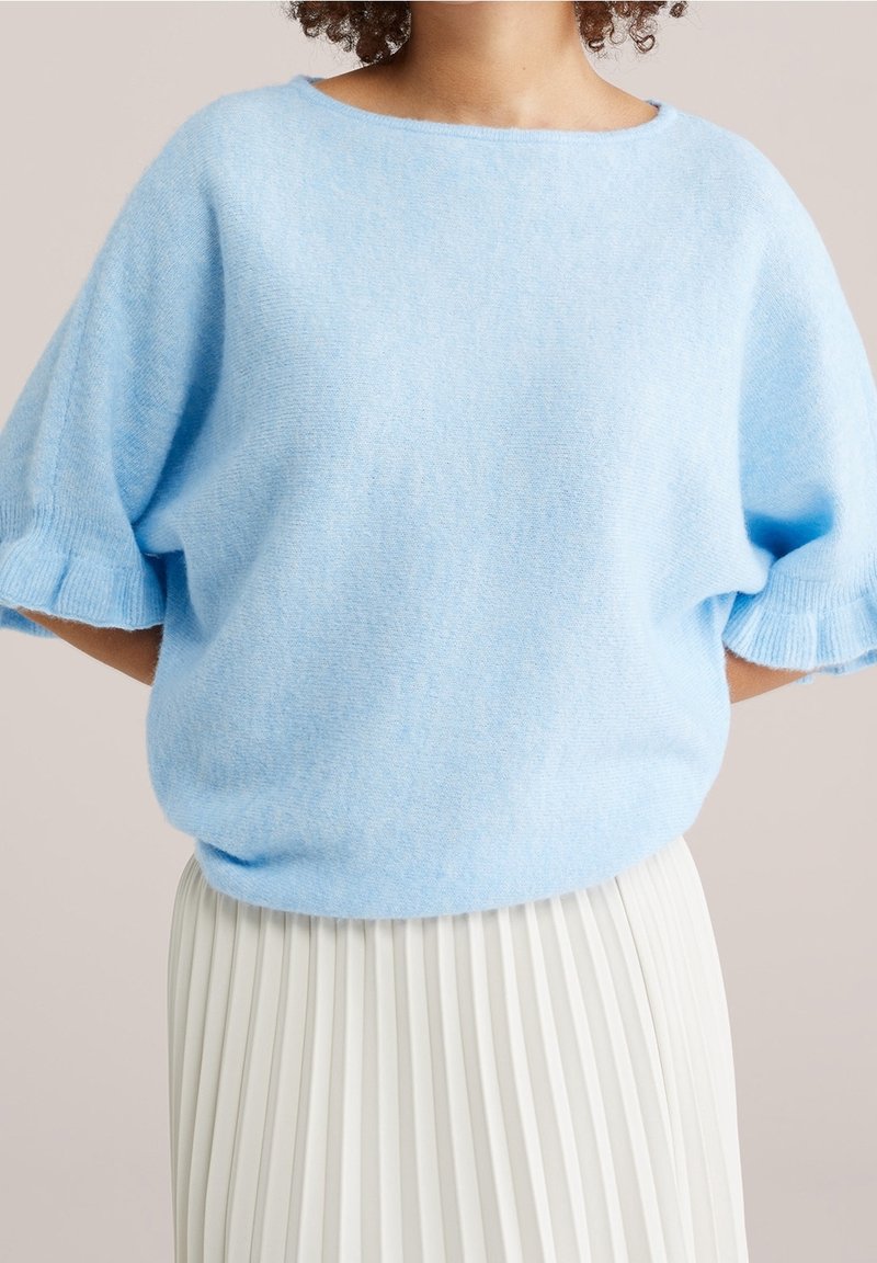 Personne portant un pull bleu clair à manches courtes avec des manches à volants et une jupe plissée blanche sur un fond neutre.