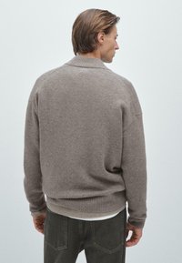 Brązowy sweter z dzianiny z prążkowanym dołem i mankietami, luźny krój oraz subtelna tekstura. Noszony z białą warstwą pod spodem.