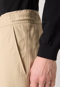 Pantalons en tissu beige avec une ceinture élastique et des poches latérales. Le nom de la marque "HUGO" est embossé sur la ceinture. Un haut noir à manches longues est visible.