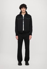 Black cropped jacket with turquoise stitching, button closure, and side pockets, paired with black joggers, worn with a graphic T-shirt.