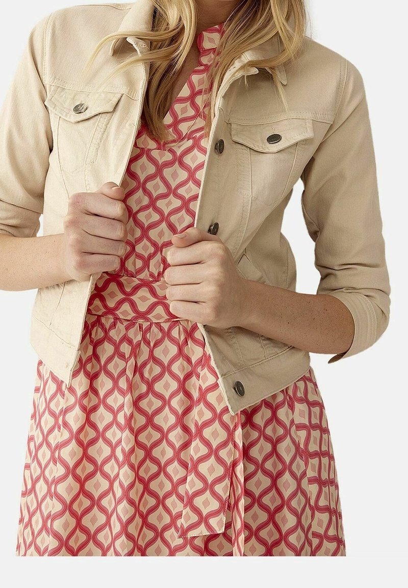 Chaqueta de mezclilla beige clara con botones en el frente y mangas cortas, usada sobre un vestido rojo y rosa con un patrón de diseño geométrico.