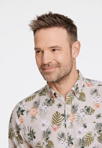 Camicia a maniche corte con bottoni di un beige chiaro, decorata con motivi floreali e foglie verdi e arancioni. Presenta un colletto classico e un dettaglio con taschino sul petto.