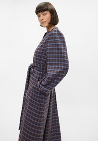 Lang rutete kjole i brunt, blått og hvitt. Har belte i midjen, lange ermer og sidelommer. Laget av et mykt, strukturert stoff.