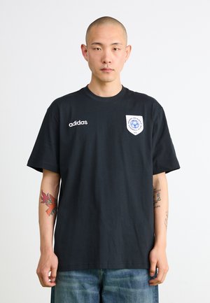 Sort bomulds t-shirt med korte ærmer, med hvid "adidas" logo og et rundt patch-design på venstre side. Casual pasform, blød tekstur.